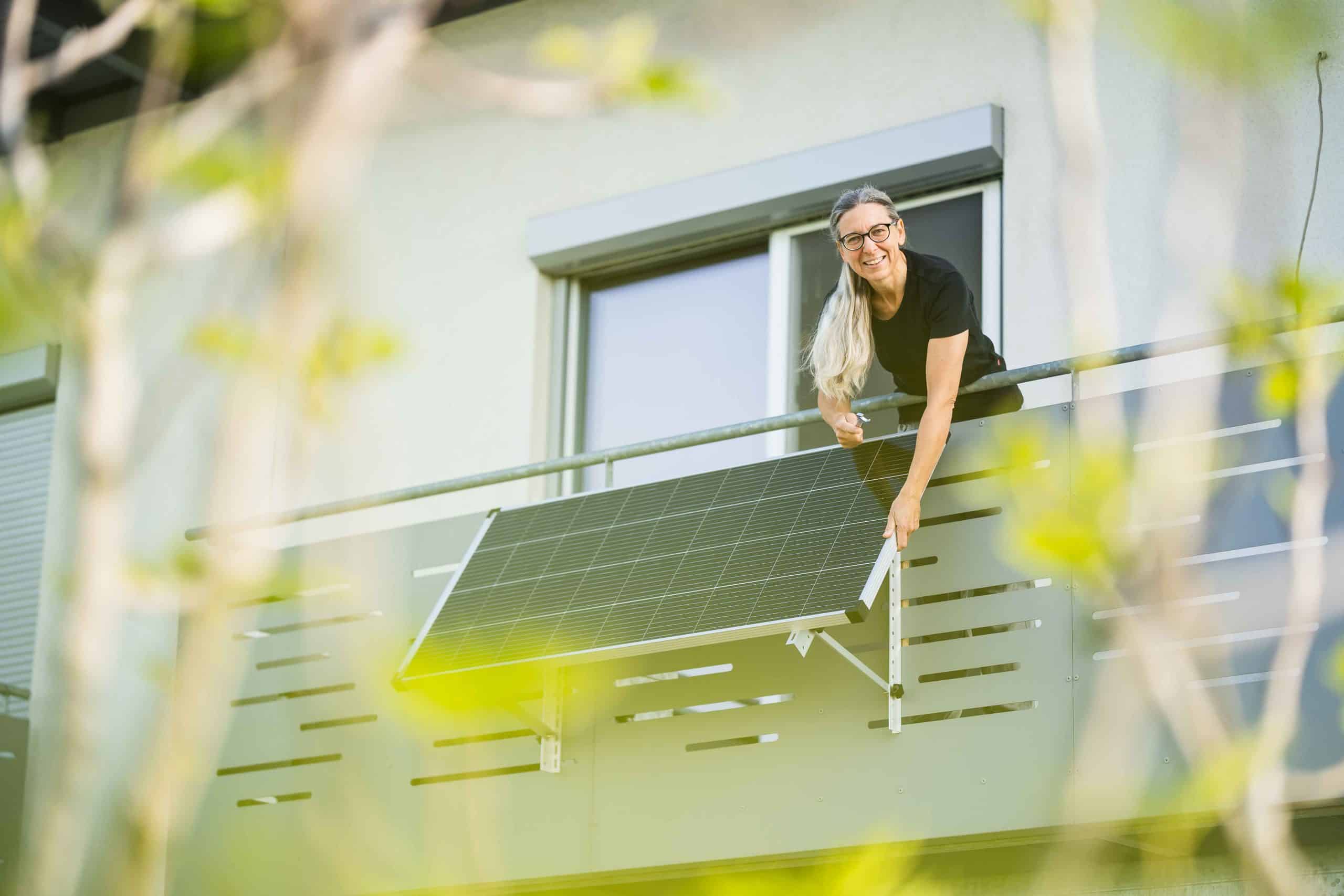 Eine Frau schaut lächelnd von ihrem Balkon herab. Ihre Hände greifen zu einer daran befestigten Solaranlage.