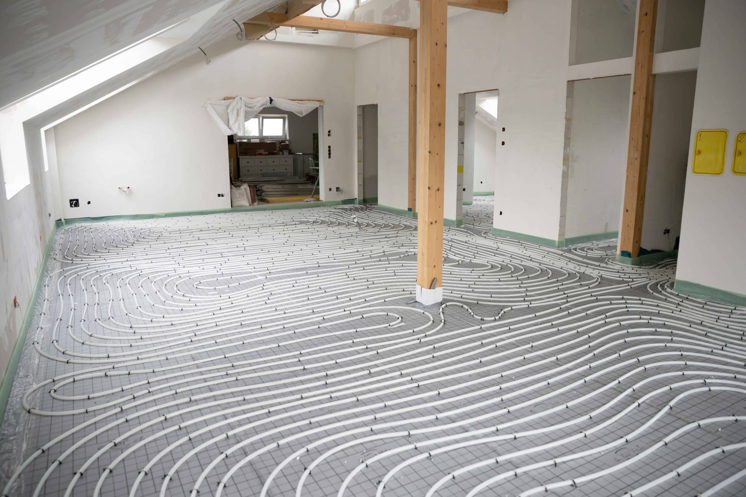 Ein großer Raum mit weißen Wänden und Tragkonstruktionen aus Holz. Weiße Schläuche für eine Fußbodenheizung sind am Boden verlegt.
