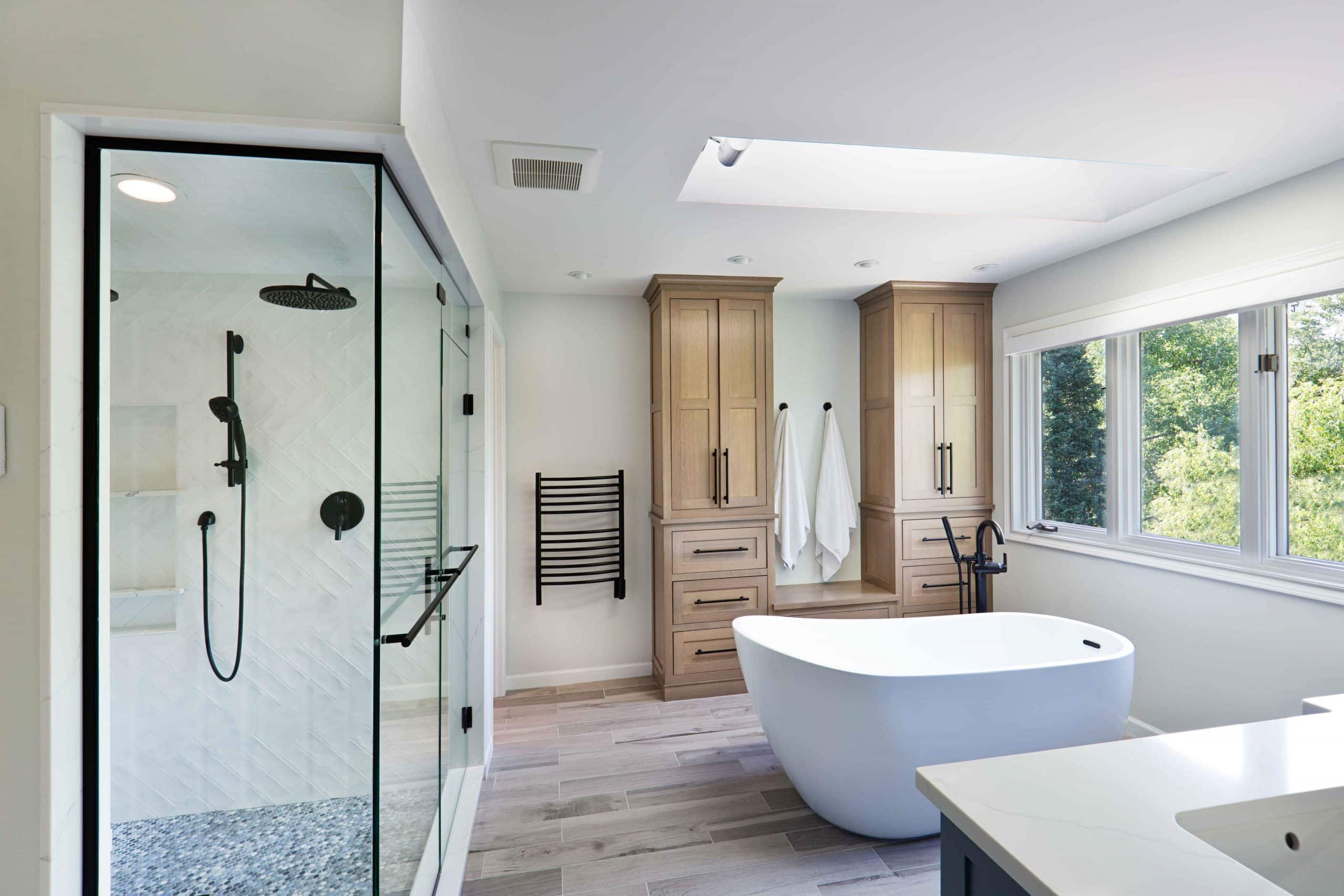 Ein Bad mit weißen Wänden und Fenstern. Darin befinden sich eine weiße Badewanne, eine schwarze Dusche mit Handbrause & Regendusche, sowie ein großer, zweigeteilter Schrank aus Holz im Hintergrund.